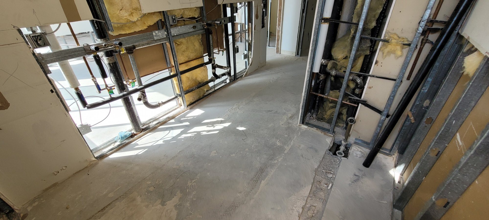 Hall walls opened to steel studs and plumbing, with a trench cut in the slab at a drain.